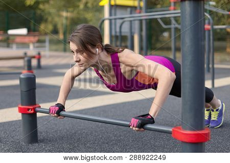 The Girl In A Bright Sportswear Pushes Up In Outdoor Sportground. White Earphones, Protective Gloves