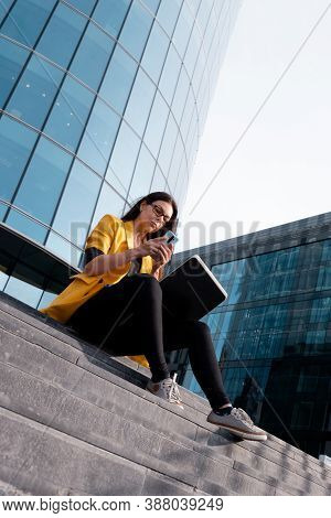 Freelancer With A Laptop Sat On The Steps Near The Business Center, A Young Woman In A Yellow Jacket