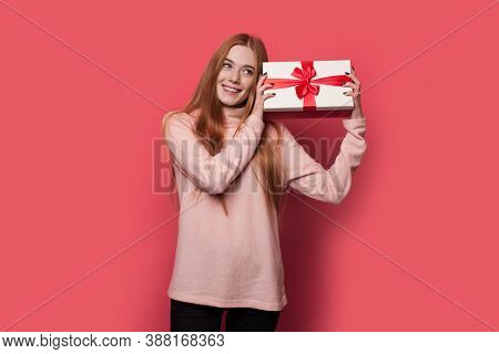 Red Haired Woman With Freckles Is Shaking A Present And Smile On A Red Wall At Studio
