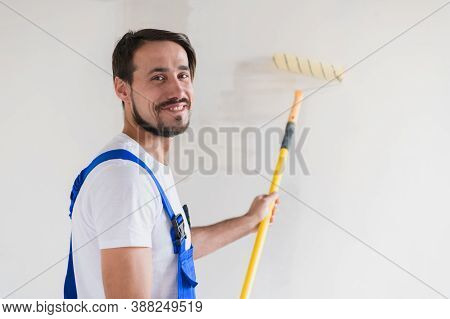 A Bearded Repaiman In Work Clothes Paints The Wall With A Roller. He Smiles Friendly And Poses For T