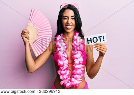 Beautiful hispanic woman wearing bikini and handfan holding hot word sticking tongue out happy with funny expression. 