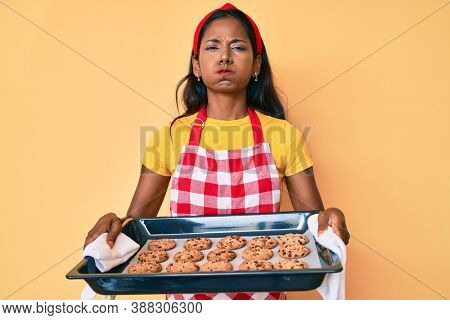 Young indian girl wearing baker uniform holding homemade cookies puffing cheeks with funny face. mouth inflated with air, catching air. 