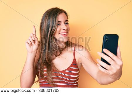 Beautiful young caucasian woman taking a selfie photo with smartphone smiling with an idea or question pointing finger with happy face, number one 