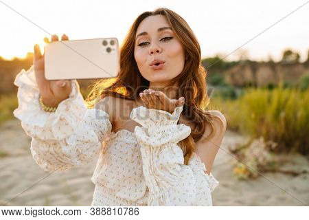 Young caucasian attractive brunette woman smiling and taking selfie phoro on cellphone outdoors