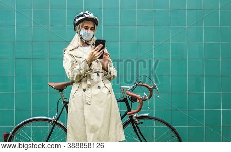 Woman in protective maskin, in helmet using her smartphone device. Female go to work on his sport retro bike. Girl standing on green background.