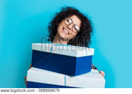 Caucasian Woman With Curly Hair And Glasses Is Giving A Present At The Camera Smiling On A Blue Stud
