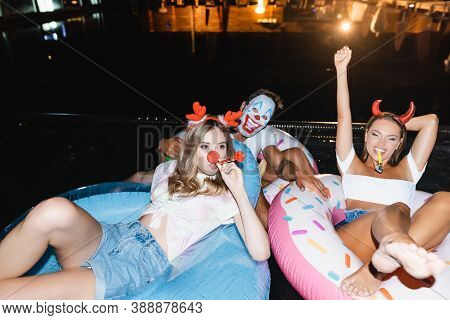 Selective Focus Of Friends With Blow Ticklers On Swim Rings In Pool At Night