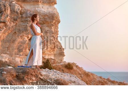 Cute Lady In White Dress Posing On Rock. Sunset Over Sea And Nature. Contemplation And Self Absorbed