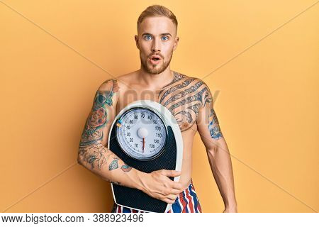Young caucasian man holding weight machine to balance weight loss scared and amazed with open mouth for surprise, disbelief face 
