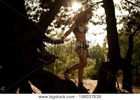 Beautiful little girl in costume of nymph dryad with floral head wreath stands on tree roots against sun in magical fairytale forest.