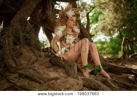 Beautiful little girl in image of nymph dryad with floral head wreath sits on tree roots in magical fairytale forest .