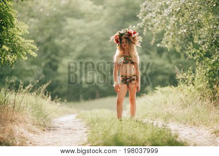 Beautiful little girl in image of nymph dryad with floral head wreath stands in forest road. Close up.