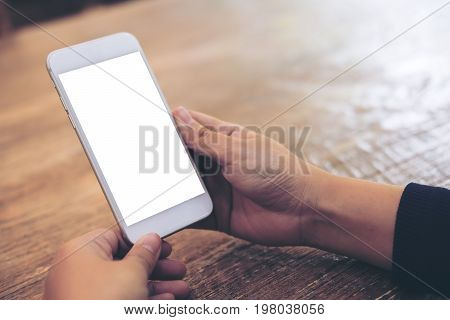 Mockup image of woman's hands holding and using white mobile phone with blank screen on wooden table background