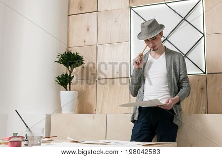 Designer thoughtfuly stands looking at paper. Young man considers on work results of designer sketch