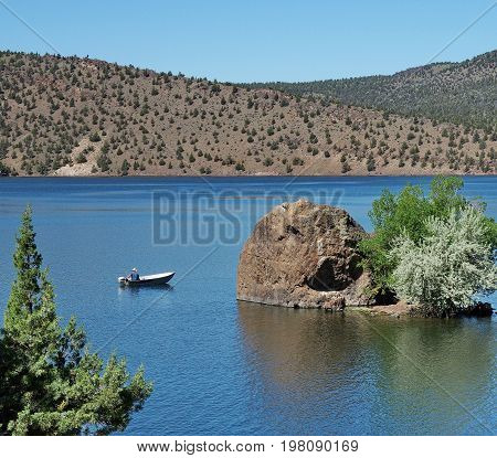 A fisherman in a small aluminum boat trolls for fish past a little boulder island with trees in Prineville Reservoir in Central Oregon on a sunny summer morning.