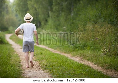 Back view on kid boy in a straw hat walking on a countryside road and playing with football ball. Child outdoors on a sunny summer day. Lifestyle vacations concept.