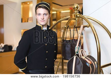 Smiling man in bellboy uniform standing with luggage cart and looking at the camera