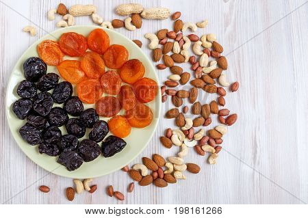 On a light table a plate of dried apricots and prunes. Near it are scattered nuts. A mixture of nuts consists of peeled and peanuts almonds and cashews.