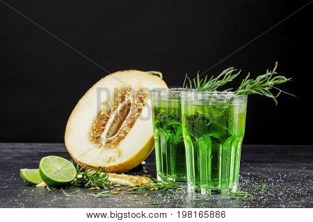 Two beautiful glasses of alcoholic drink with ice and tarragon leaves and delicious cut melon on a black background. Refreshing and fruity alcoholic summer beverages. Fresh melon and cold cocktails.