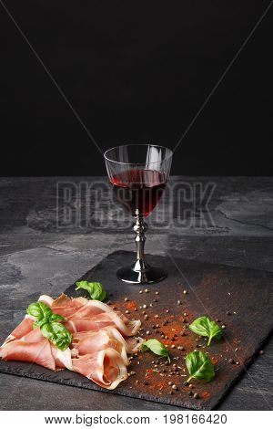 Vertical position of a wineglass filled red wine, salted and raw balyk, spicy basil on a black background. Prosciutto and semi sweet wine. A glass of wine and a pile of cut ham on a black background. Copy space.