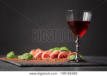 A wineglass filled red wine, salted and raw balyk, spicy basil on a black background. Prosciutto and semi sweet wine. A glass of wine and a pile of cut ham on a black background. Copy space.