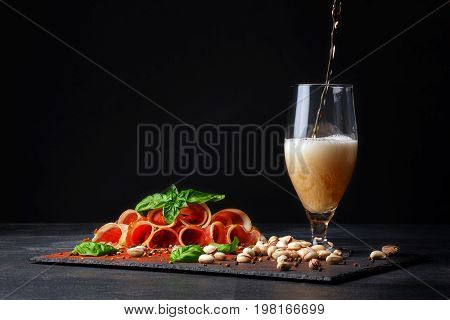 A tall and huge glass filled with golden cold beer and salted pistachios, cut twisted pieces of prosciutto on a black background. Copy space. Pistachios and a plate of sliced balyk pieces.