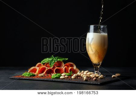 A tall and huge glass filled with golden cold beer and salted pistachios, cut twisted pieces of prosciutto on a black background. Copy space. Pistachios and a plate of sliced balyk pieces.