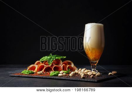 A tall and huge glass filled with golden cold beer and salted pistachios, cut twisted pieces of prosciutto on a black background. Copy space. Pistachios and a plate of sliced balyk pieces.
