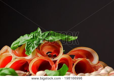 A plate of perfectly sliced prosciutto with green leaves of basil and pepper on a black background. Cured balyk with pistachios close-up. Sliced smoked bacon with pepper. Raw and salt meat. Copy space