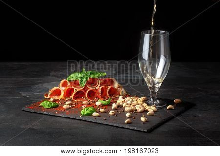 A composition of an empty glass for beer, pistachios and a plate of perfectly sliced balyk with a spinach on a black background. Prosciutto with a basil and a little bit beer in a glass. Copy space.