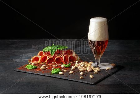 A composition of a glass of dark beer, pistachios and a plate of perfectly sliced balyk with spinach on a dark background. Cured prosciutto with a basil and a goblet of alcoholic beer with foam.