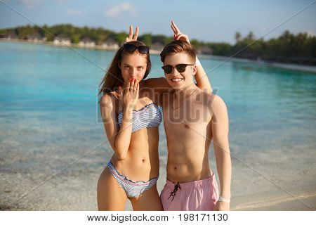 Happy young couple have fun and relax on the beach. Man and woman posing for photo and fool around. Bungalows of spa resort, palms, and blue sea on background. Family travel concept. Maldives