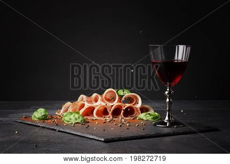 A glass of red wine and thinly sliced balyk on the saturated black background. Dry red wine, traditional Italian snacks, basil and peppercorns on the table. Copy space.