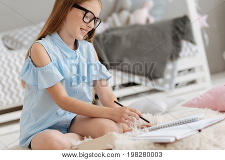 Necessary information. Side view on a female youngster in casual attire sitting on a faux fur carpet and noting something down with pencil in her hand.