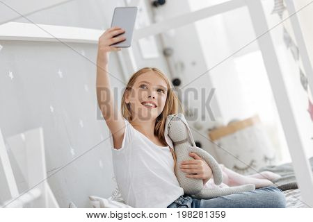 Posing together. Adorable girl grinning broadly while hugging her favorite bunny toy and taking selfies while having a rest and spending free time at home.