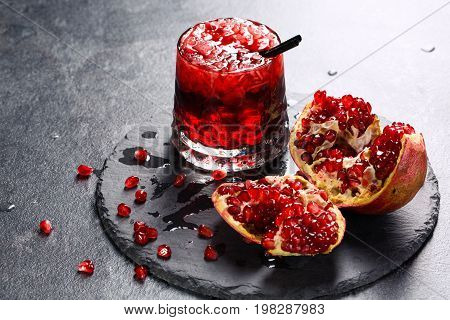 A composition of a non-alcohol beverage with garnet juice, ice and a straw. A cracked pomegranate and a glass of red juice on a tray on a gray background. Delicious garnet products full of vitamins.