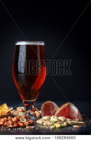 A close-up picture of a tall flass full of golden cold beer on a table. A beer with spicy snacks: salty pistachios, closed and cracked peanuts, yellow nachos chips and cut ham on a black background.