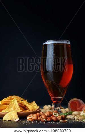 A close-up picture of a tall flass full of golden cold beer on a table. A beer with spicy snacks: salty pistachios, closed and cracked peanuts, yellow nachos chips and cut ham on a black background.