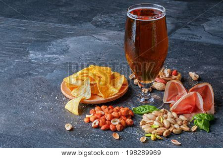 A set of tasteful and nutritious traditional snacks next to a big pint of dark beer on a black table background. Nachos chips in a plate and peanuts. Sliced ham and appetizing pistachios. Copy space.