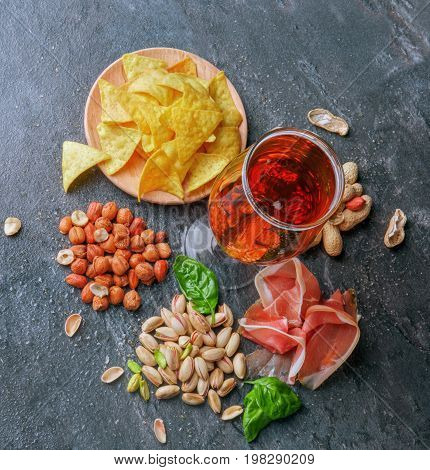 A view from above on different tasteful snacks and a tall glass of dark beer on a black table background. A plate of chips, crunchy nuts, spinach leaves and cut prosciutto. Delicious refreshing beer.