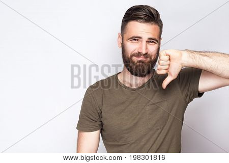 Portrait of unsatisfied bearded man with thumbs down and dark green t shirt against light gray background. studio shot.