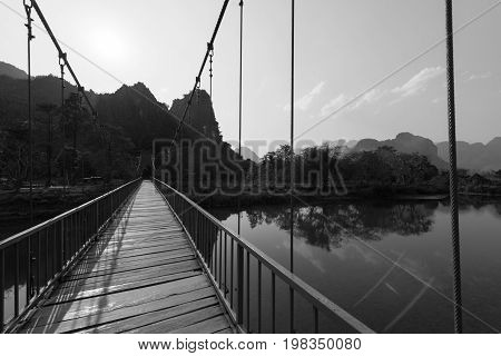 Black and white picture of orange bridge over the Nam Song River with tropical vegetation. Close to Tham Chang Cave in the city of Vang Vieng Laos.