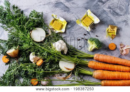Raw vegetables and bouillon cubes for cooking soup. Young carrot with haulm, broccoli, cauliflower, onion, garlic, salt pepper over gray concrete background. Top view. Dinner cooking concept