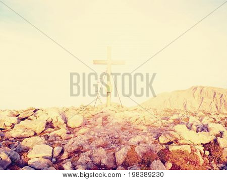 Modest Wooden Cross Raised On Rocky Alpine Mountain Summit . Sharp Rocky Peak. Gentle Clouds  In Blu