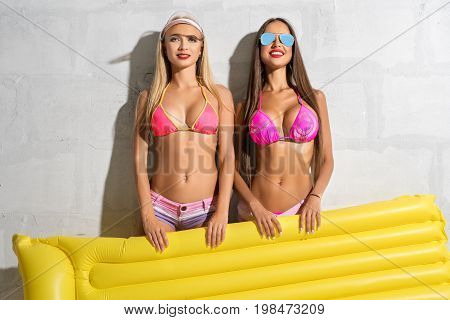 Two sexy girls wearing sunglasses and pink bicinis posing in studio near brick white wall holding yellow swimming air mattress
