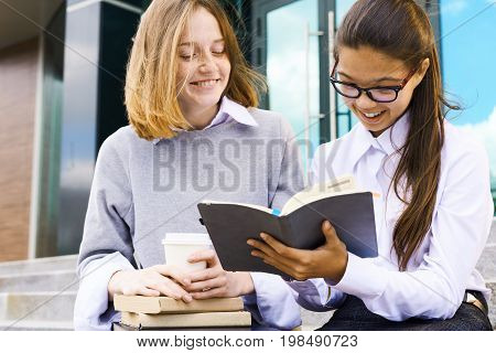 Portrait of two teenage girls studying outdoors in school yard, smiling and reading books on break