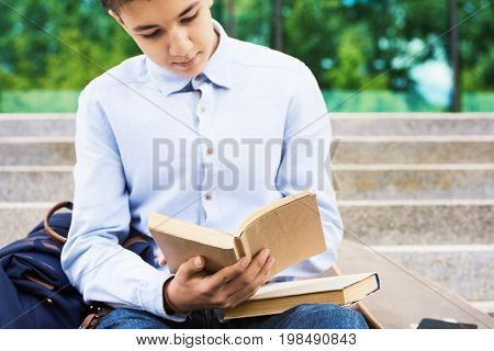 Portrait of smart teenage boy reading books for studying outdoors on stone steps of school building