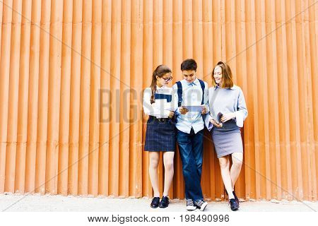 Portrait of three school children standing against bright orange wall using digital tablet