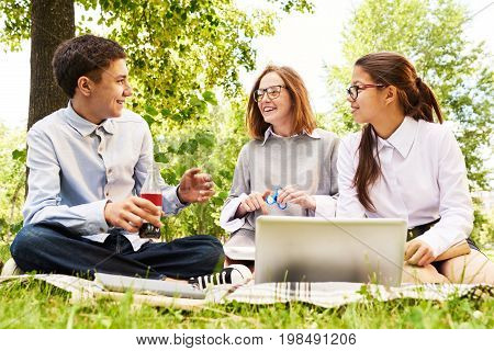 Group of modern schoolchildren enjoying picnic on green lawn, chatting and using laptop