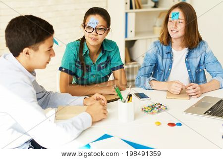 Group of teenagers playing guessing game at table in classroom with names on stickie notes